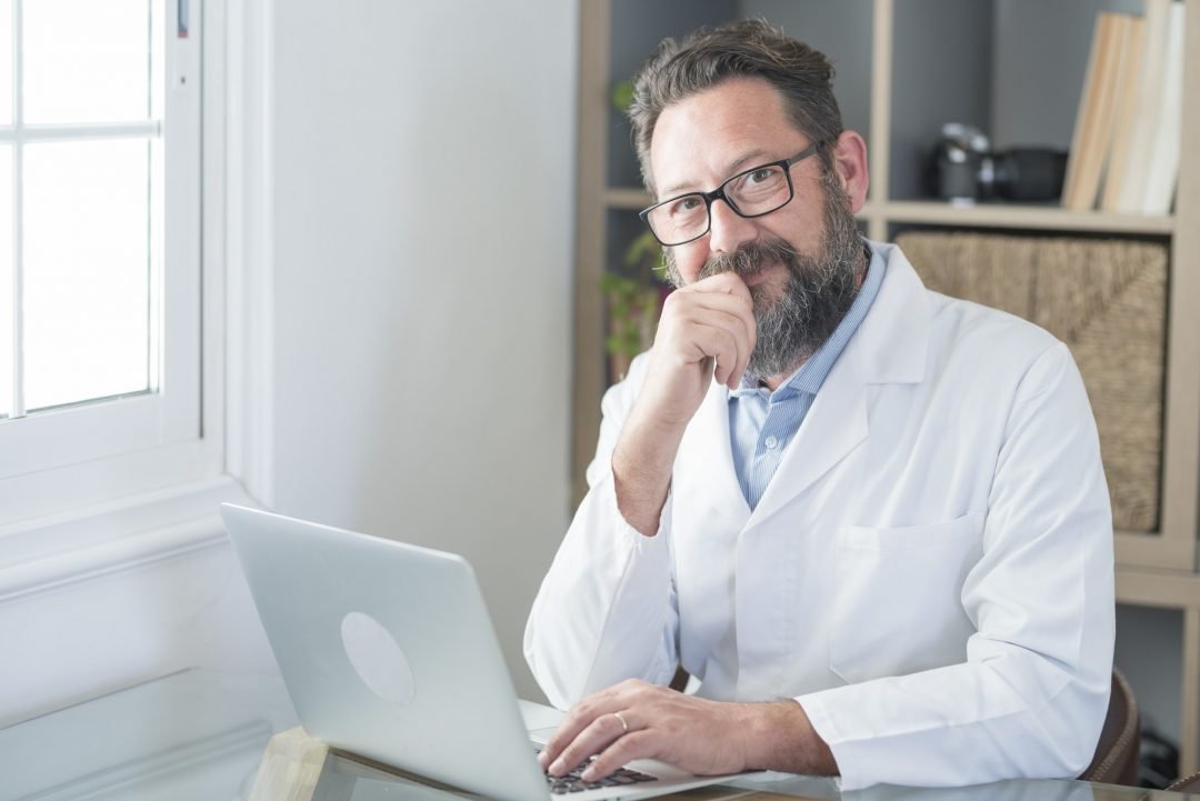 smiling-old-male-doctor-in-glasses-and-white-uniform-sit-at-desk-in-hospital-work-on-laptop.jpg